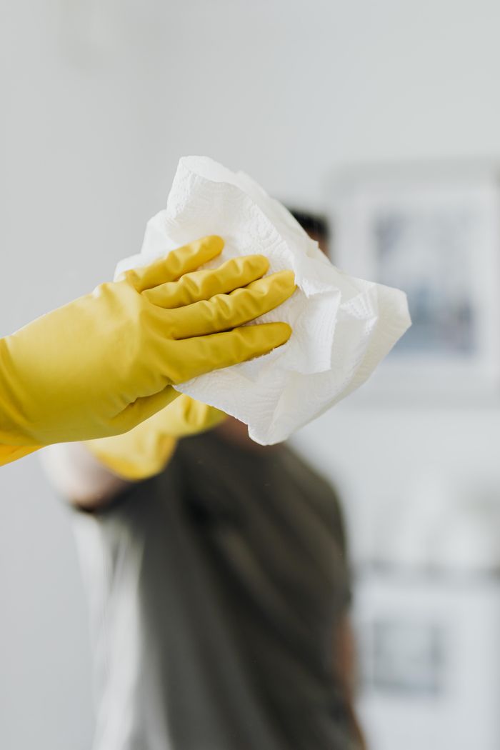 Un homme anonyme portant des gants en latex jaunes et un t-shirt foncé essuie un miroir dans une salle de bain avec une serviette en papier